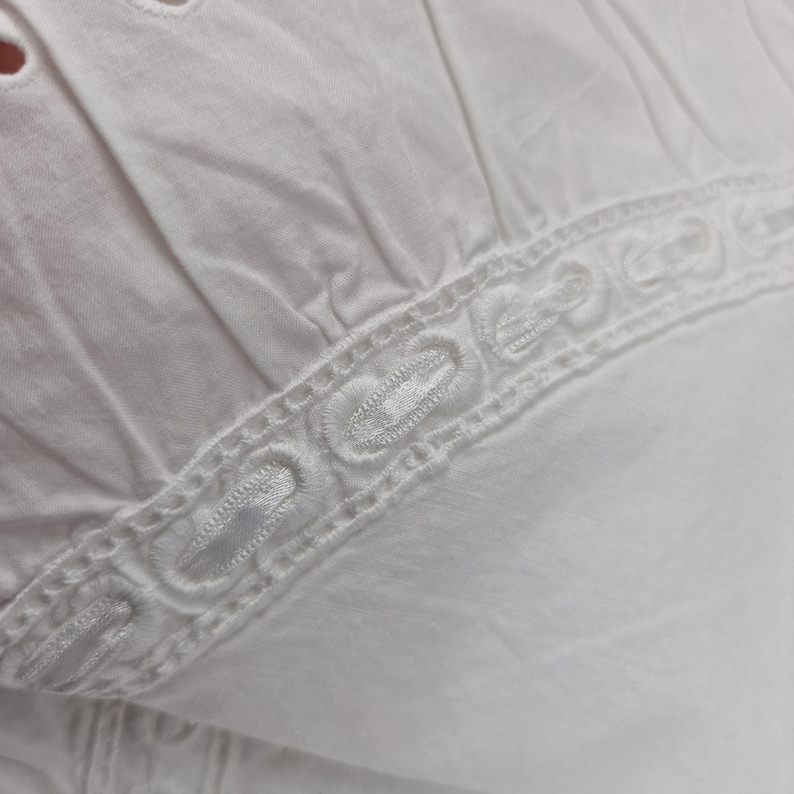May include: Close-up of white fabric with eyelet details and a decorative embroidered border. The fabric appears to be cotton or a similar lightweight material, with a slightly textured surface. The design suggests a garment or textile.