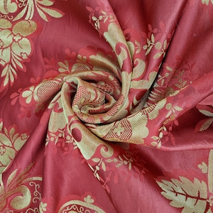 May include: A close-up of a swirled, red fabric with a gold floral pattern. The fabric has a slight sheen and a textured appearance. The gold pattern includes leaves and floral designs, creating a luxurious look. The fabric is likely used for upholstery or drapery.