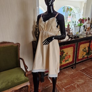 May include: A pale yellow slip dress featuring a V-neckline with white lace trim, thin pink shoulder straps, and a matching white lace hem. The garment has a gathered bodice with a small central bow detail and falls to a knee-length silhouette.