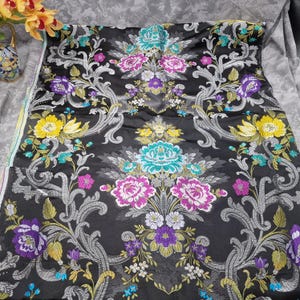May include: Black fabric with a floral pattern in shades of pink, purple, yellow, and teal. The design features large flowers and scrolling gray accents. The fabric is likely used for upholstery or decorative purposes.