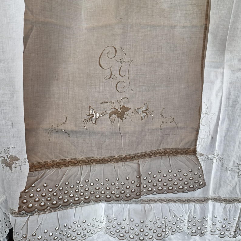 May include: A vintage beige linen textile with embroidered details. Features a monogram, floral and butterfly motifs, and a scalloped edge. The textile has a ruffled trim and is framed by white fabric with embroidered accents.