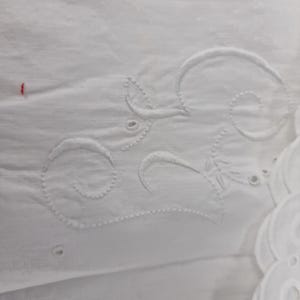 May include: Close-up of white fabric with embroidered floral and swirl designs. The embroidery is detailed and creates a textured effect on the fabric. The fabric has a soft, delicate appearance, with a few small holes.