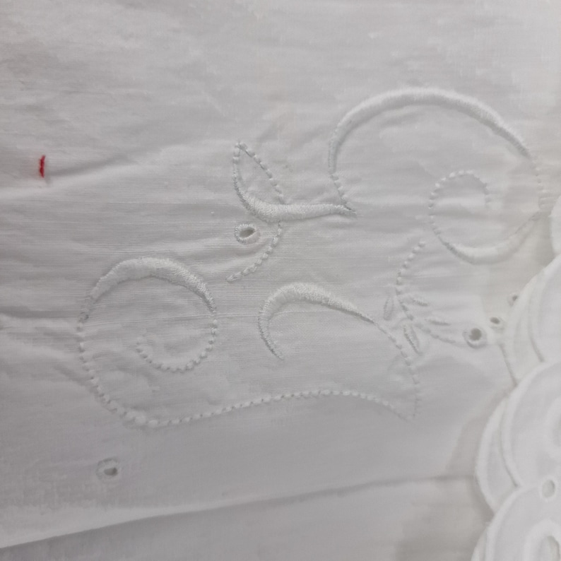 May include: Close-up of white fabric with embroidered floral and swirl designs. The embroidery is detailed and creates a textured effect on the fabric. The fabric has a soft, delicate appearance, with a few small holes.