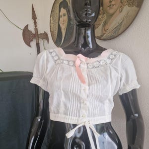 May include: A white short-sleeved blouse featuring a square neckline with intricate white lace trim, vertical pleats along the front, a central row of small buttons, and a light pink ribbon tie at the collar with a white tie at the hem.
