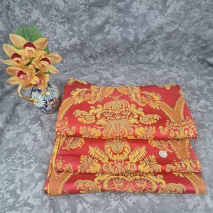 May include: Folded red fabric with a gold damask pattern. The fabric is folded in three layers. A small, decorative vase with yellow flowers and a blue and white floral design is in the background.