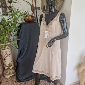 Op de afbeelding: Een beige mouwloze onderjurk met een V-hals, voorzien van gedetailleerde kanten afwerking langs het lijfje en een bijpassende kanten rand aan de zoom. Het kledingstuk is voorzien van een wit rechthoekig productlabel aan de voorzijde.