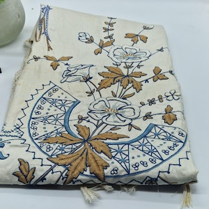 Puede incluir: Un tejido de color blanco roto plegado con intrincados bordados en tonos azules y bronceados. El diseño muestra un gran arreglo floral con pétalos y hojas detallados, complementado con un patrón de borde geométrico en forma de abanico.