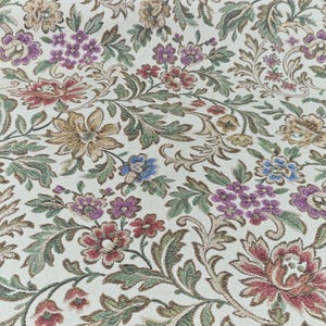 May include: Close-up of a floral fabric with a cream background. The pattern features detailed flowers in shades of red, purple, blue, and gold, with green leaves and stems. The fabric has a woven texture.