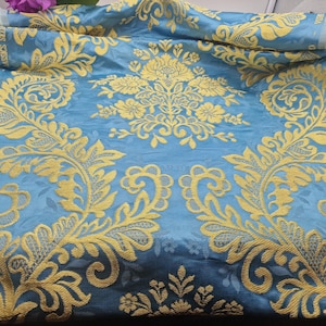 May include: A close-up of a folded fabric with a teal background and a gold floral pattern. The pattern features large, ornate floral and leaf designs. The fabric has a slight sheen, and the gold embroidery stands out against the teal.
