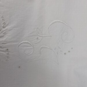 May include: Close-up of white fabric with embroidered floral and scrollwork designs. The embroidery is in a matching white thread, creating a subtle, textured pattern. The design includes leaves, circles, and curved lines.