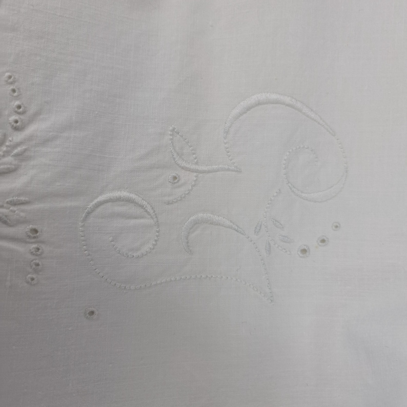 May include: Close-up of white fabric with embroidered floral and scrollwork designs. The embroidery is in a matching white thread, creating a subtle, textured pattern. The design includes leaves, circles, and curved lines.