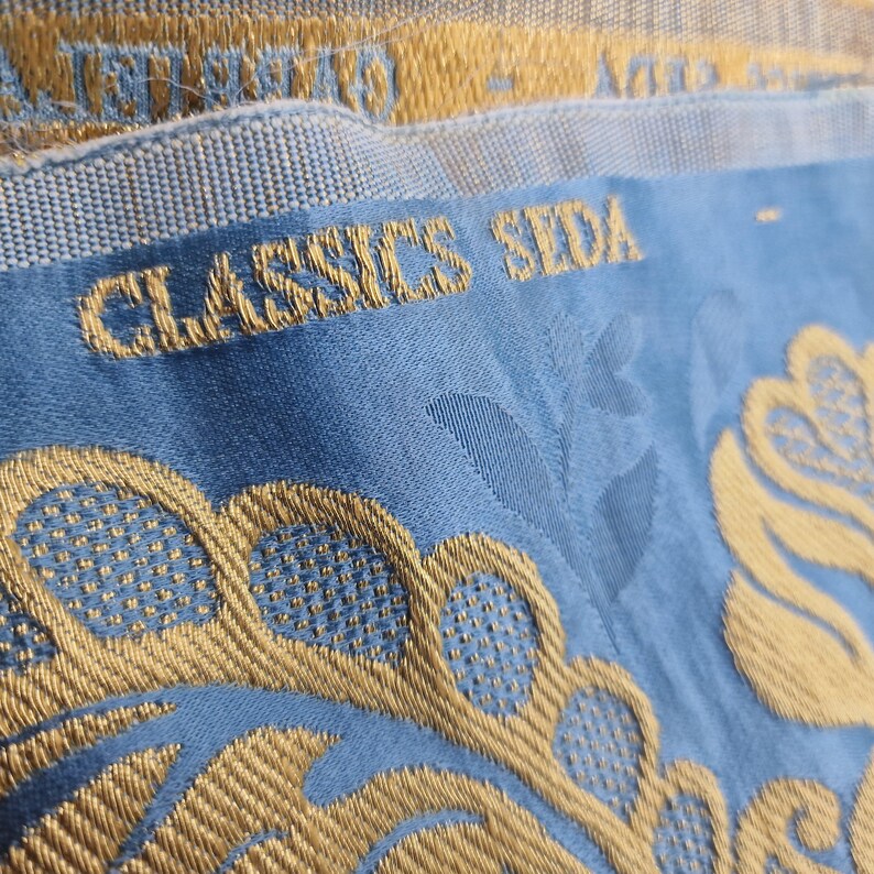 May include: Close-up of a section of fabric with the words "CLASSICS SEDA" embroidered in gold. The fabric is a light blue with a gold floral pattern. The top edge has a gold and light blue woven trim.