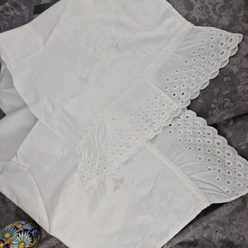 May include: Two white pillowcases with embroidered floral designs and eyelet lace trim. The pillowcases are made of a soft, white fabric and have a delicate, vintage aesthetic. The eyelet lace trim adds a touch of elegance to the bedding.