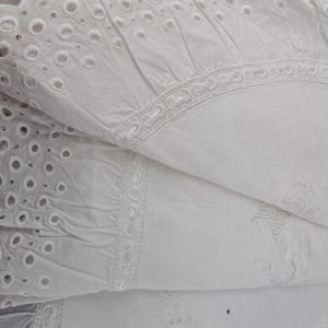 May include: Close-up of white eyelet fabric with intricate cut-out patterns and decorative embroidered borders. The fabric is folded, revealing layers of texture and detail. The eyelet design features small, evenly spaced circular holes.