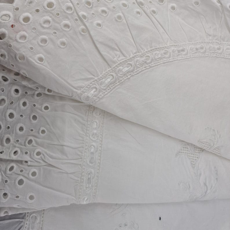 May include: Close-up of white eyelet fabric with intricate cut-out patterns and decorative embroidered borders. The fabric is folded, revealing layers of texture and detail. The eyelet design features small, evenly spaced circular holes.