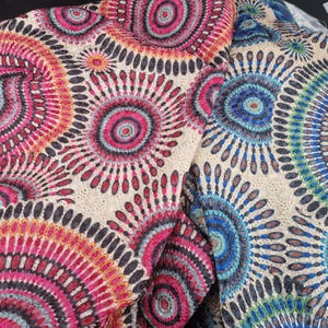 May include: Close-up of two fabric pieces with a cream background. The fabric has a repeating circular pattern in pink, red, blue, and black. The design resembles a stylized floral or mandala motif, creating a vibrant and intricate textile.