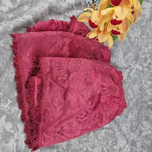 May include: A stack of deep rose-colored fabric with a floral pattern and fringed edges. The fabric is folded, revealing the intricate design. Yellow and red orchids are in the background.