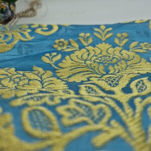 May include: Close-up of a teal fabric with a gold floral pattern. The design features large, ornate flowers and leaves. The fabric has a slight sheen, giving it a luxurious appearance. The pattern is detailed and intricate, creating a rich texture.