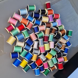 Puede incluir: Una gran colección de carretes de hilo de coser en una amplia variedad de colores vibrantes, incluyendo rojo, azul, verde, amarillo, rosa y morado. Cada carrete cuenta con un núcleo de plástico con hilo enrollado, dispuesto en un montón.