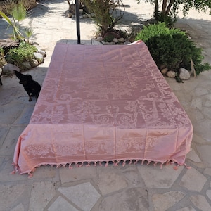 Può includere: Una tovaglia rettangolare color rosa polvere con un motivo damascato tono su tono di fiori e volute. Il tessuto ha una lucentezza sottile ed è rifinito con una frangia decorativa sul bordo inferiore, che ricade su una superficie piana.