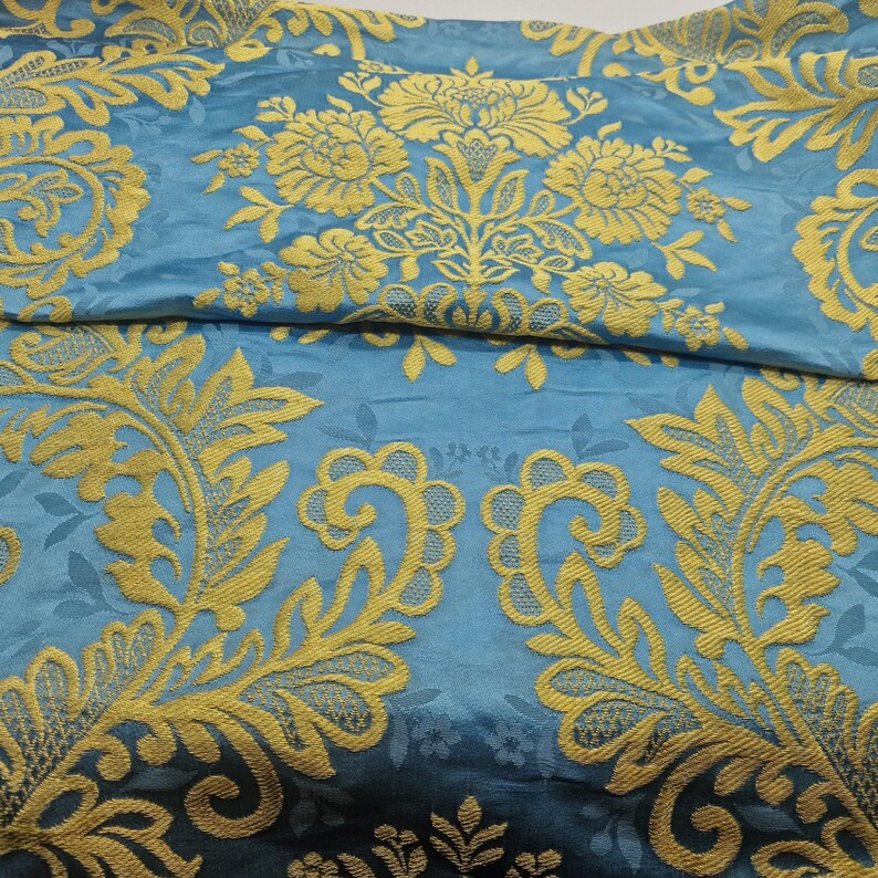 May include: A close-up shot of folded fabric. The fabric is teal blue with a gold floral pattern. The pattern features large, ornate flowers and leaves, creating a luxurious and elegant look. The fabric has a slight sheen, enhancing the visual appeal.