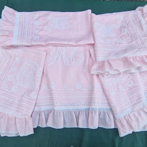 Op de afbeelding: Een set lichtroze beddengoed met witte geborduurde bloemmotieven en monograminitialen. De stukken zijn voorzien van ruches met ingewikkelde wit kantachtige details langs de randen, wat zorgt voor een samenhangende textielcollectie.