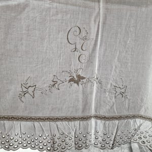 May include: A light-colored linen textile with embroidered details. The design includes floral motifs and the initials "G" and "C". The bottom edge features a scalloped eyelet trim, adding a delicate touch to the piece.