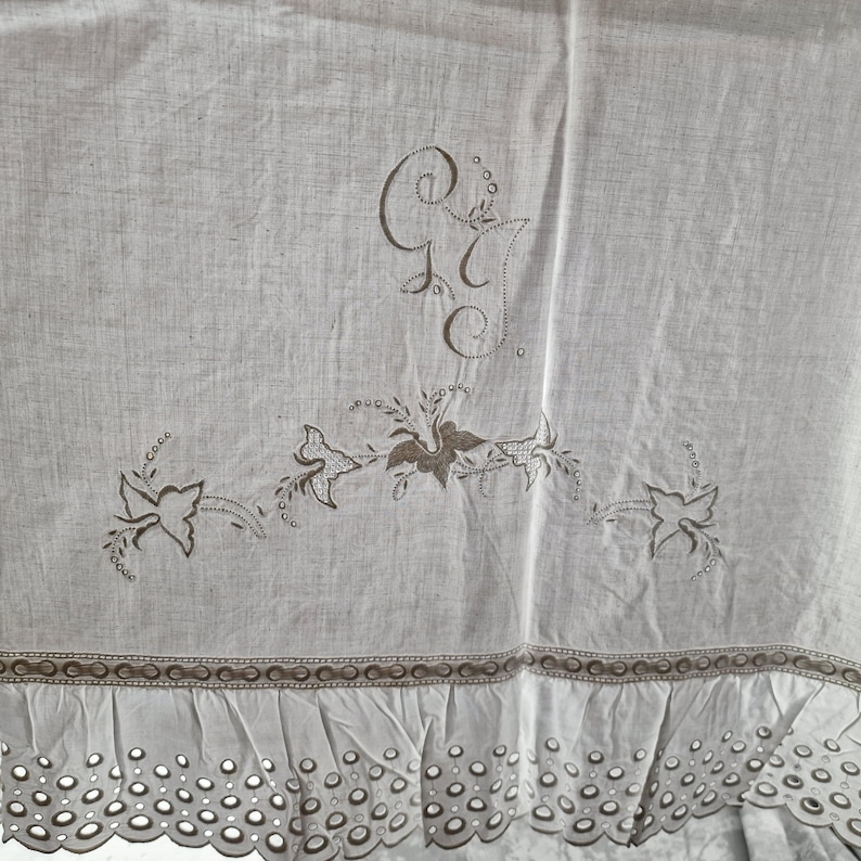 May include: A light-colored linen textile with embroidered details. The design includes floral motifs and the initials "G" and "C". The bottom edge features a scalloped eyelet trim, adding a delicate touch to the piece.