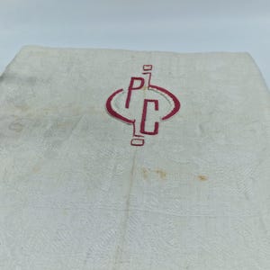 Puede incluir: Una tela textil blanca con un sutil patrón de damasco floral tejido en toda su superficie. Centrado en la tela hay un monograma bordado en hilo rojo, que muestra las letras P y C encerradas dentro de una forma geométrica redondeada y estilizada.