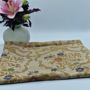 Puede incluir: Una tela beige doblada con un estampado floral en dorado, azul, rojo y verde. La tela está decorada con un diseño repetido de flores y cintas. Un pequeño jarrón redondo verde claro con lirios rosas está en el fondo.
