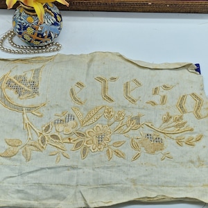 Puede incluir: Textil bordado de color crema con la palabra "ETES" y diseños florales. El bordado es de color marrón claro, con una luna creciente y patrones florales. El textil parece envejecido, con algo de decoloración.