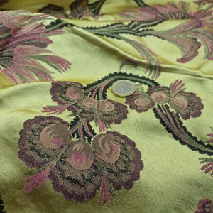 May include: Close-up of a textured fabric with a floral pattern. The fabric has a yellow-green background with a repeating design of burgundy and pink flowers and black accents. The fabric is folded, showing the texture and depth of the material.