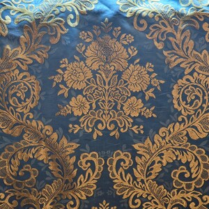 May include: Close-up of a blue fabric with an ornate gold floral pattern. The design features large, stylized flowers and swirling leaf motifs. The fabric has a subtle sheen, enhancing the gold embroidery. The overall aesthetic is elegant and decorative.