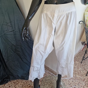 May include: White cotton-style cropped trousers featuring a smooth waistband and intricate lace detailing along the lower leg hems. The pants have a relaxed fit with a straight-leg silhouette that ends mid-calf, showcasing geometric lace inserts.