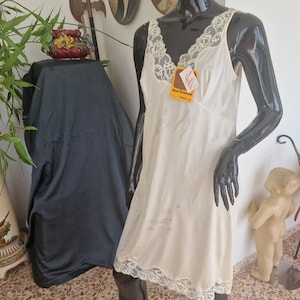 Pode incluir: Um vestido tipo combinação sem mangas na cor creme com decote em V adornado com renda floral branca e bainha de renda a condizer. Uma pequena etiqueta retangular amarela com texto em vermelho e preto está presa na parte frontal da peça.