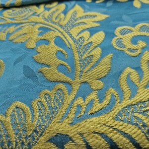 May include: Close-up of a teal fabric with a gold floral pattern. The design features stylized leaves and swirling shapes, creating a textured, ornate appearance. The gold elements are raised, adding depth to the textile.