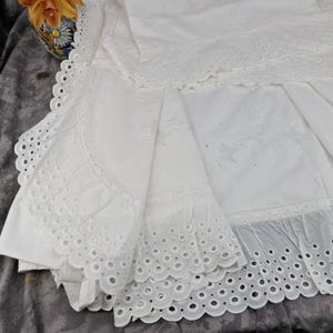 May include: White eyelet fabric with scalloped edges and intricate cut-out designs. The fabric is folded, revealing layers of detailed embroidery and a delicate, airy texture. The overall aesthetic is vintage and elegant.