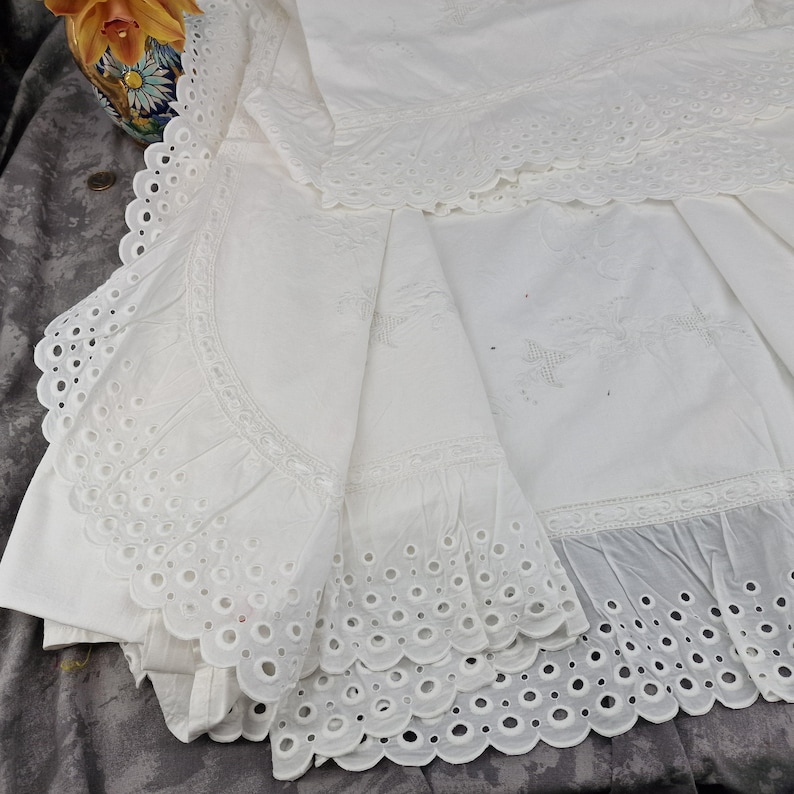 May include: White eyelet fabric with scalloped edges and intricate cut-out designs. The fabric is folded, revealing layers of detailed embroidery and a delicate, airy texture. The overall aesthetic is vintage and elegant.