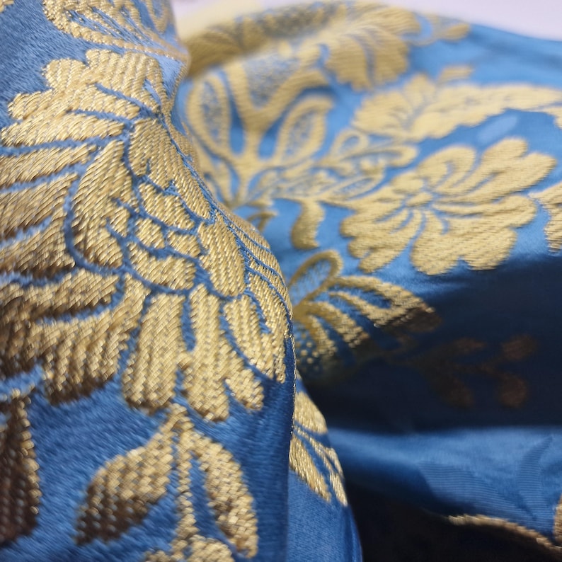 May include: Close-up of a luxurious blue fabric with gold floral embroidery. The intricate design features detailed leaves and swirling patterns, creating a rich, textured appearance. The fabric is folded, showing the depth and sheen of the material.