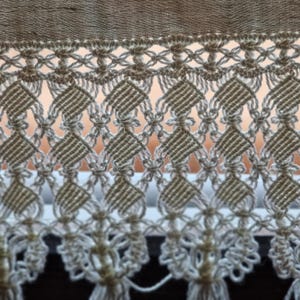 Puede incluir: Primer plano de un textil de macramé beige. El intrincado diseño presenta formas de diamante y motivos florales, con una franja en la parte inferior. El macramé se utiliza probablemente para la decoración del hogar, como una cortina o un tapiz de pared.