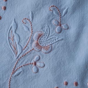Puede incluir: Una superficie de tela blanca con un delicado bordado en hilo color melocotón y blanco. El diseño muestra una flor central estilizada con detalles intrincados en los pétalos, rodeada de hojas delgadas, pequeñas flores y puntos.