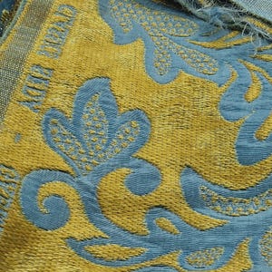 May include: Close-up of a textured fabric with a repeating floral pattern. The design features gold and teal blue colours. The fabric has a woven appearance, with the words "CHRISTIAN DIOR" visible in gold.