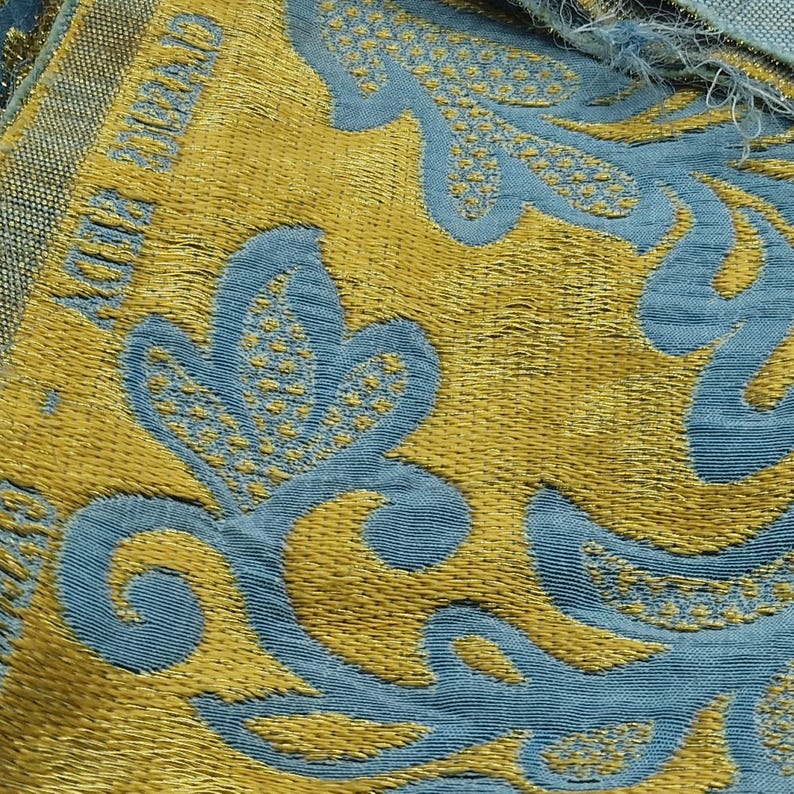 May include: Close-up of a textured fabric with a repeating floral pattern. The design features gold and teal blue colours. The fabric has a woven appearance, with the words "CHRISTIAN DIOR" visible in gold.