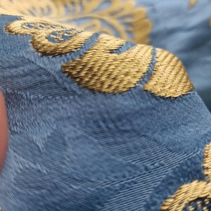 May include: Close-up of a section of fabric with a blue background and gold floral embroidery. The gold embroidery has a textured appearance, and the fabric has a slight sheen. The image highlights the intricate details of the textile.