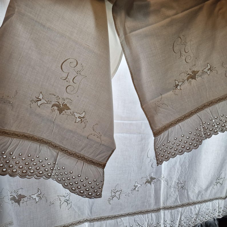 May include: Two off-white linen panels with embroidered butterflies, floral accents, and initials. The panels have a scalloped edge with small circular cutouts and a decorative border. The fabric is slightly translucent.