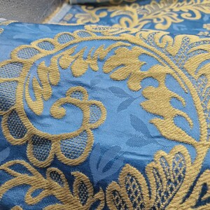 May include: Close-up of a blue fabric with a gold floral pattern. The fabric has a textured appearance with the gold design embroidered over the blue base. The design includes stylized flowers and leaves, creating a luxurious look.
