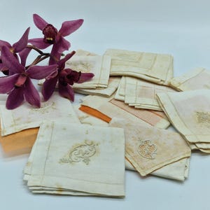 Puede incluir: Una colección de servilletas de lino vintage en varios tonos crema y beige, algunas con diseños florales o monogramas bordados. Algunas servilletas tienen detalles en naranja. Una ramita de orquídeas moradas añade un toque de color.