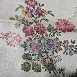 Könnte beinhalten: Ein weißer Stoff mit einem gewebten Blumenmuster und einem zentralen Strauß aus rosa, blauen, gelben und violetten Blüten. Das Design umfasst grüne Blätter und Stängel, einen dezent strukturierten Hintergrund und eine spitzenartige Bordüre.