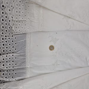 May include: Close-up of white fabric with intricate details. The fabric features eyelet lace, ruffled edges, and delicate embroidered floral patterns. A coin is placed on the fabric for scale, highlighting the fine craftsmanship and texture of the textile.