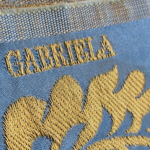 May include: Close-up of a textile with the name "GABRIELA" embroidered in gold thread. The text is on a light blue fabric, with gold floral embroidery at the bottom. The top of the textile has a gold and white woven border.